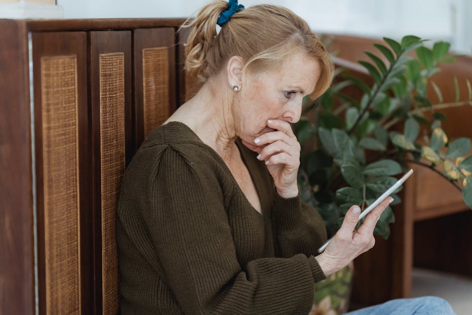Side view of surprised elderly female with cellphone covering mouth against cabinet and plant at home