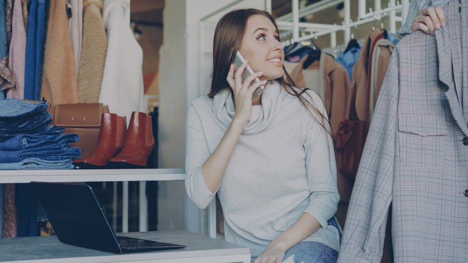 Woman in clothing store chatting on phone, surrounded by fashion items.